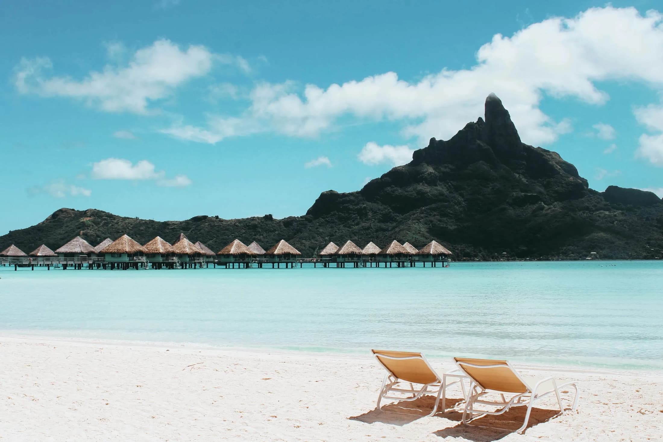 Two loungers on a white beach facing overwater bungalows and Mount Otemanu in Bora Bora
